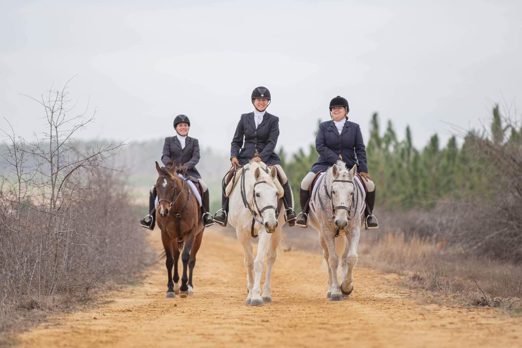 Three hunt riders on horses, walking dirt path.